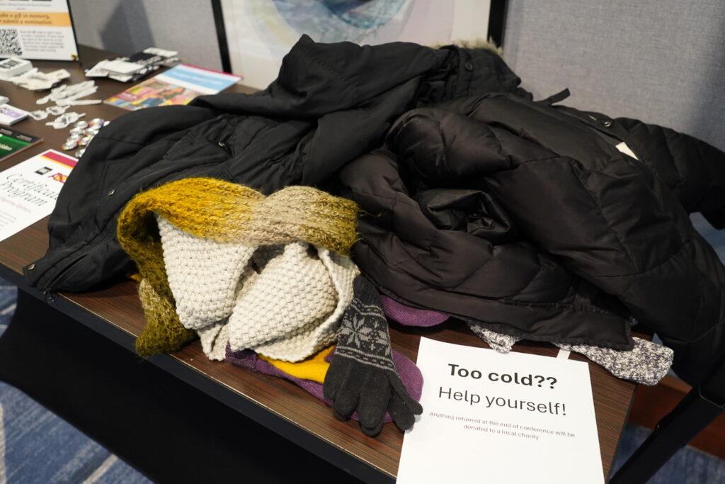A pile of coats and scarves, with a sign "Too cold?? Help yourself!"