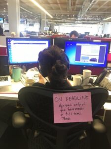 A photo of Susan Carroll from the back while she sitting at her desk with headphones. On the back of her chair is a sign that says "on deadline; approach only if you have snacks or 9-1-1 tapes."