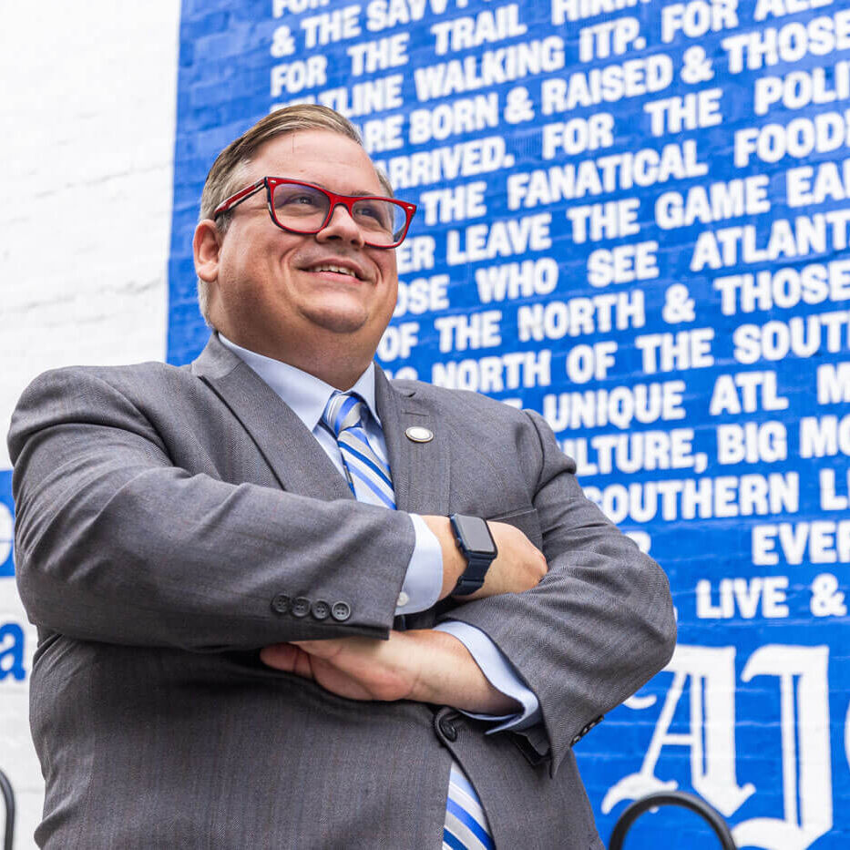 Portrait of Charles Minshew with arms crossed standing in front of a blue wall with the Atlanta Journal Constitution logo