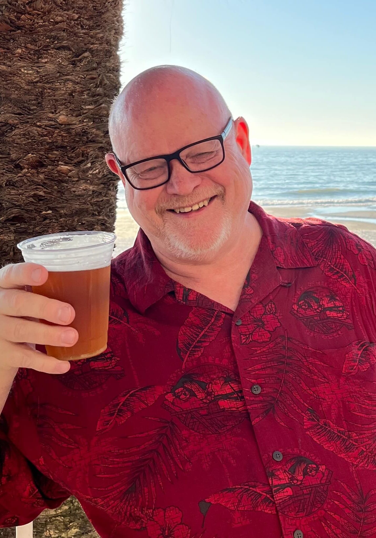 A portrait of Mark Lagerkvist holding up a beer by the beach and smiling